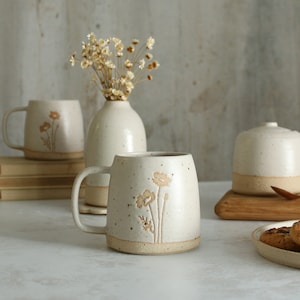 May include: Cream-coloured ceramic mug with a floral design. The mug has a rounded shape with a handle and a speckled finish. A matching vase and other ceramic pieces are also visible, creating a cohesive set.