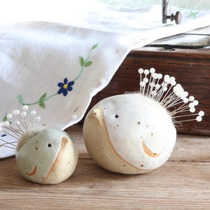 May include: Two round, ceramic bird-shaped pincushions in off-white and light gray, each featuring a small beak, dark eyes, and a cluster of silver sewing pins with white pearl heads inserted into the top to resemble a crest of feathers.