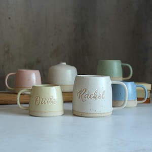 May include: A selection of handcrafted ceramic mugs in pastel shades such as pink, green, and blue. Some mugs are inscribed with names like "Rachel" and "Ottilie". The mugs have a speckled, rustic finish, ideal for tea or coffee.