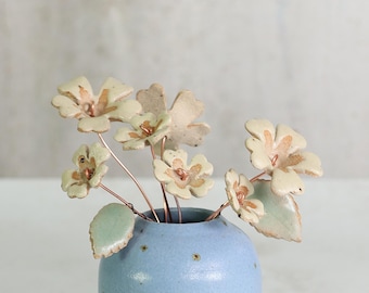 May include: A small, light blue ceramic vase with a cluster of handmade ceramic flowers. The flowers are off-white with copper wire stems and light green leaves. The vase has a speckled design.