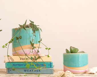 May include: Two ceramic planters with a turquoise glaze and a pink and yellow stripe. The larger planter is filled with a green plant and sits on top of a stack of books. The smaller planter is filled with a succulent and sits on a wooden surface with seashells.