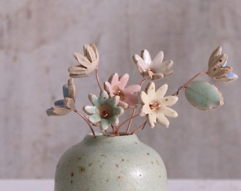 May include: A small, speckled green ceramic vase with a bouquet of pastel ceramic flowers. The flowers are in shades of pink, blue, and green.
