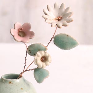 Op de afbeelding: Een keramische bloemstuk met roze, witte en groene bloemen en bladeren. De bloemen zijn bevestigd aan koperen draadstengels en zijn gerangschikt in een kleine groene vaas.