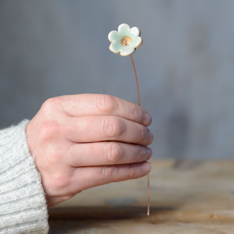 May include: A small, pale mint green ceramic flower with five rounded petals and a central hole, attached to a thin, straight copper-colored metal wire stem. The flower has a smooth, glossy finish and a slightly irregular, hand-formed shape.