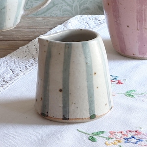 May include: A small ceramic creamer pitcher with a speckled off-white glaze and vertical sage green stripes. The pitcher features a simple pouring spout and a tapered cylindrical shape, showcasing a natural clay base and subtle brown speckles.