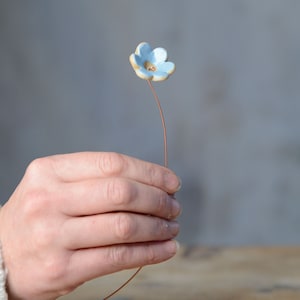 May include: A small, light blue ceramic flower with five rounded petals and a central copper-colored detail, attached to a thin, flexible copper-toned wire stem. The flower has a smooth, glossy finish and a slightly irregular, hand-crafted appearance.