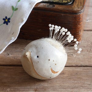 May include: A round, off-white ceramic pincushion shaped like a bird with a speckled finish and a small beak. The top features a soft, light-colored fabric insert filled with numerous silver sewing pins topped with small, round white beads.