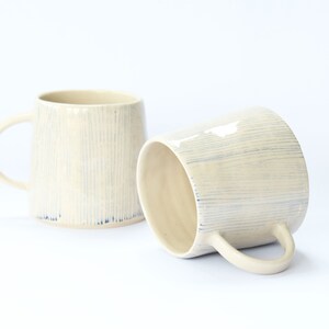 May include: Two ceramic mugs with a white glaze and blue stripes. The mugs have a rounded shape and a handle.