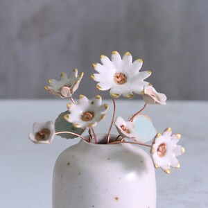 May include: A small, speckled white ceramic vase holds a delicate arrangement of handmade ceramic flowers. The white daisy-like blooms have gold-painted edges and centers, with copper wire stems and pale green leaves.