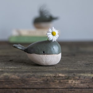 Puede incluir: Una pequeña figura de ballena de cerámica con la parte superior gris oscuro lisa y la parte inferior beige claro texturizada. La ballena tiene un pequeño ojo negro y un orificio en el lomo que sostiene una margarita blanca con centro amarillo.