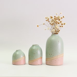 May include: Three ceramic vases in varying sizes, each with a two-tone design. The top half is a speckled green, the middle a tan stripe, and the bottom pink. The largest vase holds dried flowers.