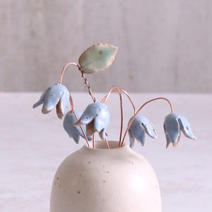 Può includere: Un piccolo vaso di ceramica bianca con una finitura macchiettata contiene un bouquet di delicati fiori di ceramica blu con steli di filo di rame. I fiori hanno un dettaglio a forma di foglia di colore blu-verde chiaro.