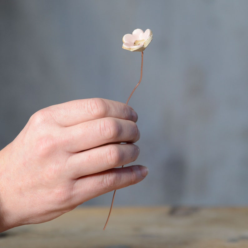 May include: A small, pale pink ceramic flower attached to a thin, flexible copper-colored wire stem. The flower features five rounded petals with a textured, off-white center, creating a delicate and simple botanical ornament for decorative use.
