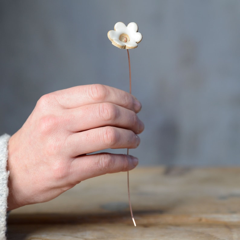 May include: A small, white, five-petaled ceramic flower attached to a thin, straight copper-colored metal wire stem. The flower has a smooth, glossy finish on the petals and a small, recessed center point where the wire connects to the ceramic.