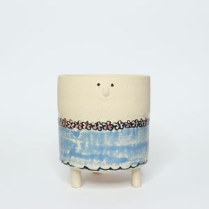 Op de afbeelding: Een witte keramische plantenpot met een blauw en roze bloemenontwerp en een glimlachend gezicht. De pot heeft drie korte pootjes.