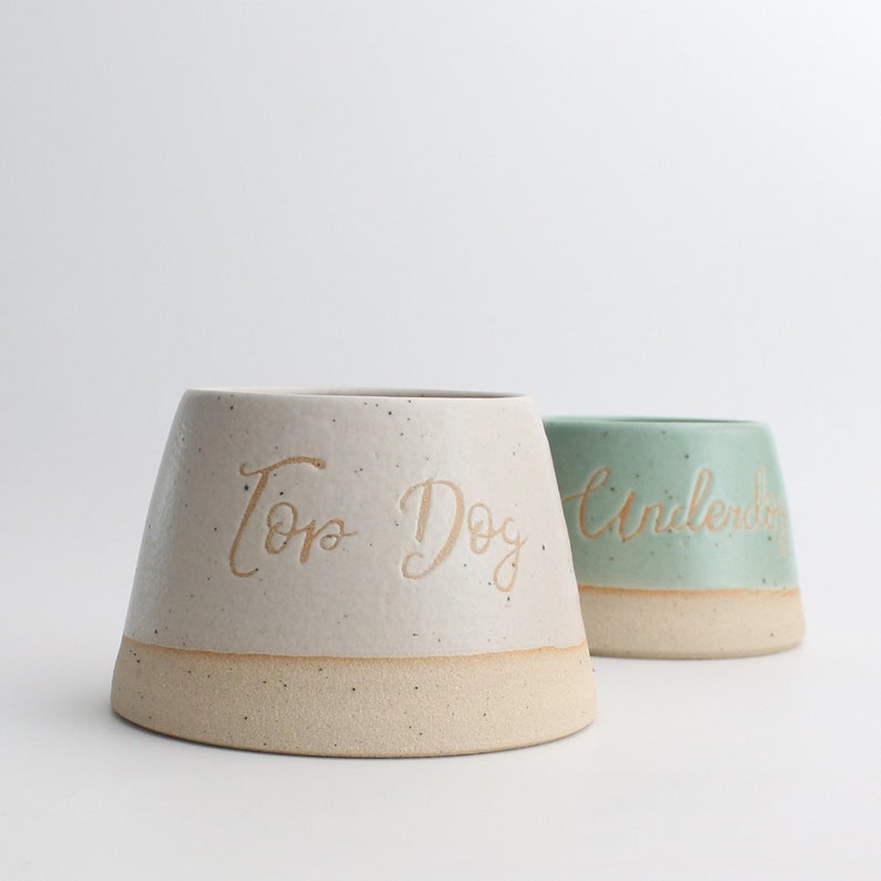 May include: Two ceramic dog bowls, one white with the text "Top Dog" and one light green with the text "Underdog".