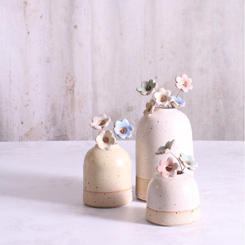 May include: Three ceramic vases with a speckled cream finish and a smooth base. Each vase features a narrow opening holding delicate, hand-sculpted ceramic flowers in soft pastel shades of pink, blue, and white, attached to thin metal stems.