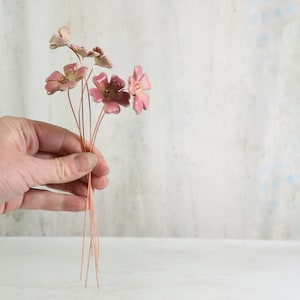 Peut inclure: Un petit bouquet de fleurs en céramique rose avec des tiges en fil de cuivre. Les fleurs ont une finition brillante et sont tenues dans une main sur un fond neutre. Les fleurs ont un aspect délicat et artisanal.