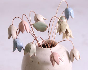 May include: A white ceramic vase with a speckled glaze holds a bouquet of pastel-colored ceramic flowers. The flowers are made to look like bell-shaped blossoms and are attached to thin copper wires.