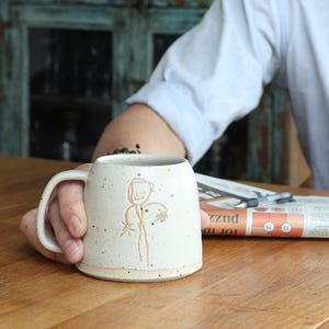 May include: A speckled off-white ceramic mug with a wide base and a curved handle. The exterior features a simple, light brown line drawing of a stick figure with a round head and outstretched arms, etched into the textured clay surface.
