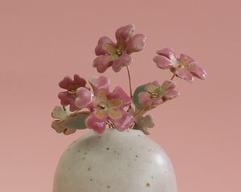 May include: A small, speckled white ceramic vase holds a delicate arrangement of pink ceramic flowers with gold centers and green leaves. The flowers are on thin copper wire stems, set against a soft pink background.
