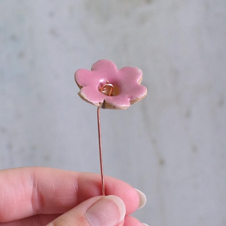 May include: A small, five-petaled pink ceramic flower attached to a thin, straight copper-colored metal wire stem. The center of the flower features a small, coiled wire detail, and the edges of the ceramic petals show a natural, unglazed texture.
