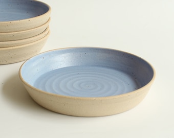 May include: A stack of shallow ceramic bowls with a light blue interior glaze featuring subtle spiral textures and dark speckles. The exterior of the bowls is a matte, unglazed beige clay finish with a visible speckled texture throughout the surface.