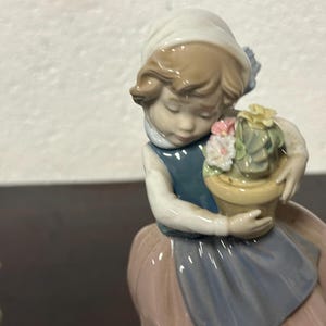 May include: A porcelain figurine of a girl holding a potted plant with pink and yellow flowers. The girl is wearing a blue dress with a white collar and a pink skirt. The figurine is in a vintage style.