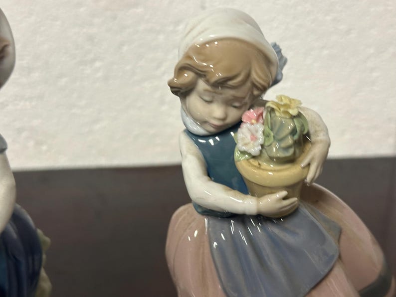 May include: A porcelain figurine of a girl holding a potted plant with pink and yellow flowers. The girl is wearing a blue dress with a white collar and a pink skirt. The figurine is in a vintage style.
