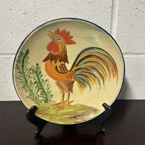 Può includere: Un piatto in ceramica con un design dipinto a mano di un gallo con una coda colorata. Il gallo è in piedi su un pezzo di erba verde. Il piatto ha un bordo blu ed è su un supporto nero.