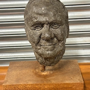 May include: A brown clay sculpture of a man's head on a wooden base.