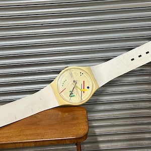May include: A large wall clock designed to resemble a wristwatch with a long white strap, a cream-coloured casing, and a round face featuring a white background, thin black hour markers, and abstract red, yellow, blue, and green geometric shapes.