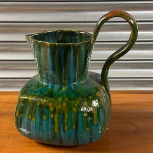 Op de afbeelding: Een groene en blauwe keramische kan met een uniek glazuurpatroon. De kan heeft een brede basis en een rond lichaam met een handvat.