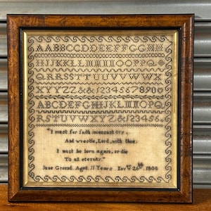 以下が含まれることがあります： 暗い木製のフレームに入ったアンティークのサンプラー。サンプラーには、刺繍された文字、数字、詩が描かれています。「I must be born again, or die」というフレーズが含まれています。サンプラーの日付は1844年11月26日です。