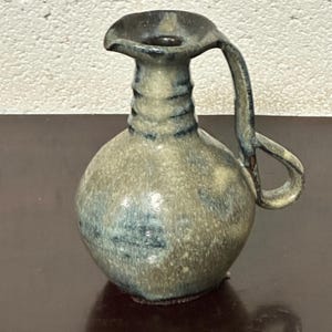 Puede incluir: Jarra de cerámica pequeña, hecha a mano, con cuerpo redondeado y asa curva. La jarra tiene un pico y está esmaltada en tonos azules, beiges y marrones. La cerámica tiene un aspecto rústico y artesanal.