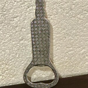 May include: A silver bottle opener with a diamond-plate pattern. The opener is shaped like a bottle with a circular opening at the bottom. The background is a textured, off-white wall.