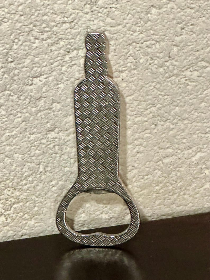 May include: A silver bottle opener with a diamond-plate pattern. The opener is shaped like a bottle with a circular opening at the bottom. The background is a textured, off-white wall.