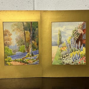 Può includere: Due dipinti ad acquerello incorniciati. Il dipinto a sinistra raffigura una scena di foresta con alberi e un ruscello blu. Il dipinto a destra mostra un giardino con fiori e un edificio. Entrambi i dipinti sono in cornici dorate.