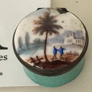 May include: A small, round, blue and white porcelain trinket box with a silver lid. The lid features a hand-painted scene of a palm tree, two figures, and a building in the background.