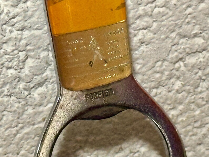 May include: Close-up of a vintage bottle opener with a silver-colored metal handle and a yellow-orange top. The word "FOREIGN" is stamped on the handle. The opener is likely used for opening bottles.