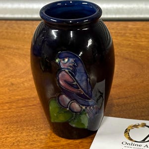 May include: A small, dark blue ceramic vase with a glossy finish. The vase features a detailed illustration of a bird in shades of blue, purple, and green. The bird is perched on a branch with green leaves. The vase has a dark blue interior.