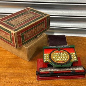 May include: A vintage Simplex typewriter and its original box. The typewriter is red, green, and black, with a circular keyboard. The box is brown with red and green accents and the words "Simplex Typewriter".