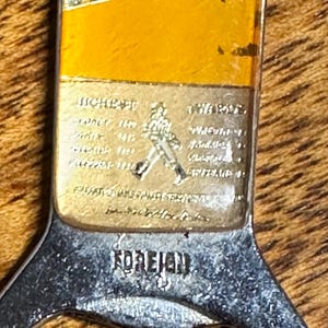 May include: Vintage bottle opener with a silver handle and a rectangular top featuring a gold and red label. The label includes text and a logo of a man walking. The word "FOREIGN" is stamped on the handle.