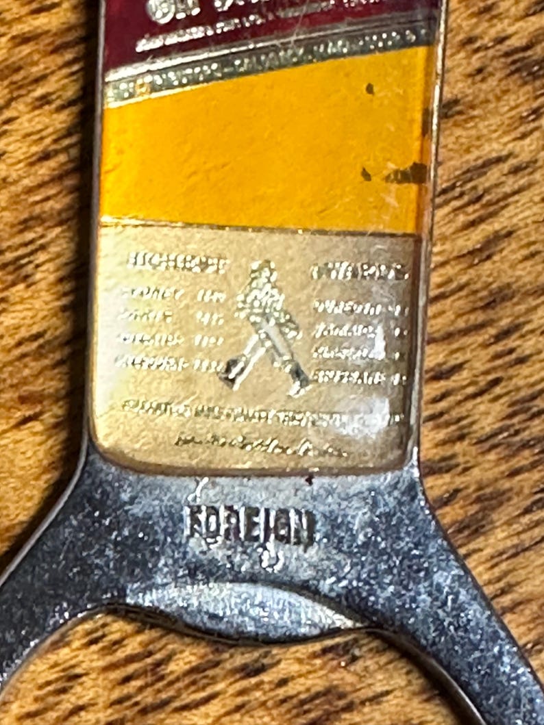 May include: Vintage bottle opener with a silver handle and a rectangular top featuring a gold and red label. The label includes text and a logo of a man walking. The word "FOREIGN" is stamped on the handle.