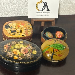 Op de afbeelding: Vier handgeschilderde houten dozen met ingewikkelde ontwerpen. Een doos heeft een gouden en zwart ontwerp met een knuffelend stel, een andere heeft een bloemenpatroon, een derde heeft een vogel op een tak en de laatste doos bevat twee kleinere dozen met bloemenpatronen.