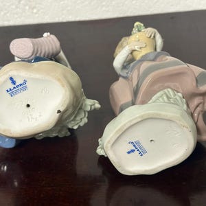 May include: Two porcelain figurines, one in blue and white and the other in pink and white, with the Lladro logo on the bottom of each.