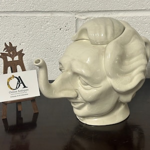 Puede incluir: Una tetera de cerámica blanca con forma de cabeza humana y un pico en forma de trompa de elefante. La tetera tiene una tapa con forma de pelo.