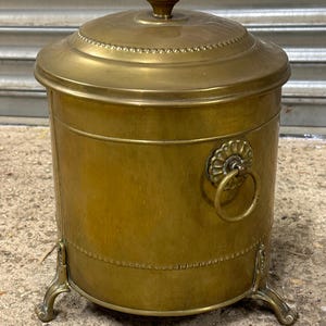May include: A vintage brass container with a lid and a decorative handle. The container has a cylindrical shape, a lid with a small knob, and rests on three ornate feet. The brass has a warm, aged patina.