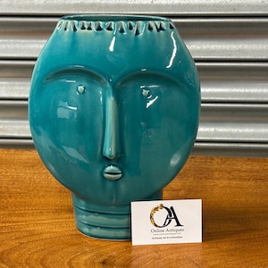 May include: A teal ceramic vase with a face design. The vase has a rounded shape with facial features including eyes, a nose, and a mouth. The vase is on a wooden surface. A business card with the logo "Online Antiques" is in front of the vase.