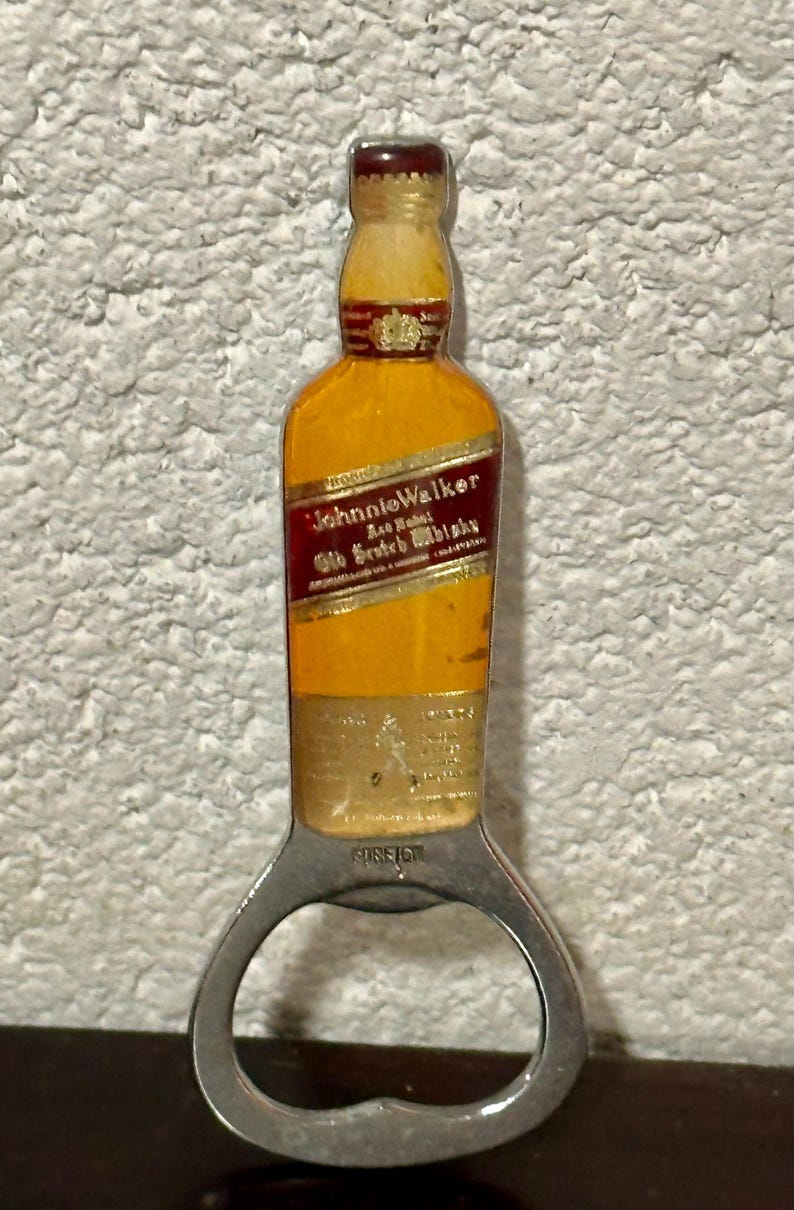 May include: Bottle opener shaped like a Johnnie Walker Scotch whisky bottle. The opener is yellow and red with the brand name and text. The bottom part is a silver bottle opener with the word "FOREIGN" printed on it.
